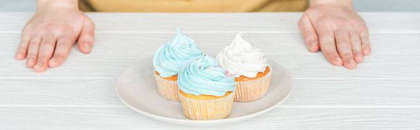 panoramic shot of woman near plate with delicious cupcakes on white table - Фото, зображення