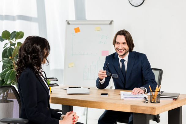 happy recruiter holding glasses and looking at brunette woman in office  - Fotoğraf, Görsel
