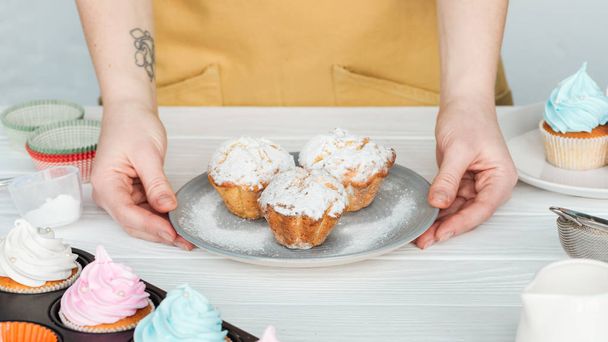Cropped view of woman holding plate with cupcakes with powdered sugar on grey - Photo, Image