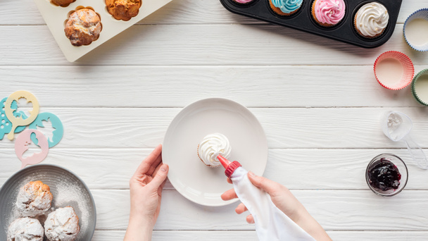 Cropped view of woman decorating cupcake with icing bag on table with cupcake trays - Photo, Image