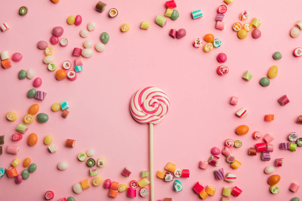 Top uitzicht van heerlijke Swirl Lollipop op houten stok met verspreide karamelsnoepjes rond op roze achtergrond - Foto, afbeelding