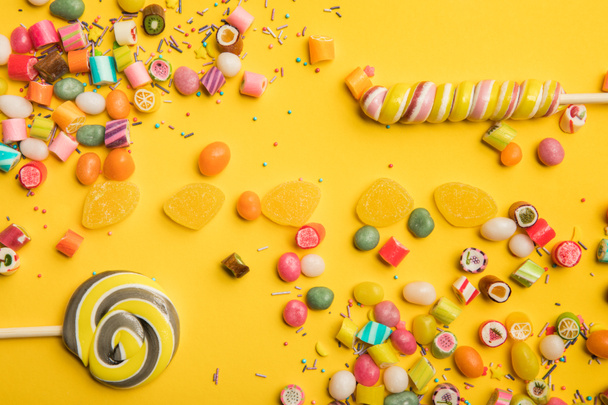 top view of delicious multicolored candies, jellies, sprinkles and lollipops on yellow background - Photo, Image