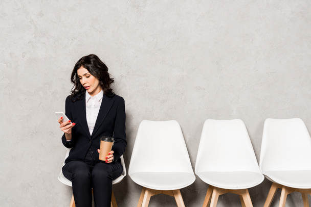 donna attraente utilizzando smartphone mentre tenendo tazza di carta e seduto sulla sedia
 - Foto, immagini