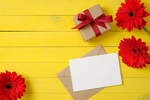 Blank paper card, vintage envelope, red daisy flowers, gift box on yellow wooden background. Women desk. Congratulation or celebration concept. Flat lay, top view, overhead. - 写真・画像