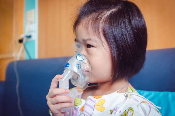 Sad asian child holds a mask vapor inhaler for treatment of asthma. Breathing through a steam nebulizer.Illness girl admitted in hospital. - Photo, Image