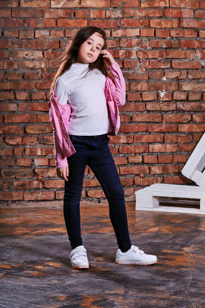 Stylish model child posing background brick wall,loft interior. Fashion girl kid hipster in casual clothes,pink jacket, white sneakers, jeans. Lifestyle, modern youth clothing. - Photo, Image