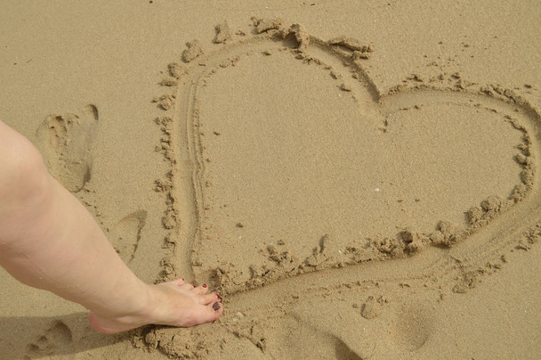 こんにちは夏、女性の足は砂浜に立っている間、濡れた砂の上にハートサインを描きます。休息、リラクゼーション、トップビューの概念 - 写真・画像