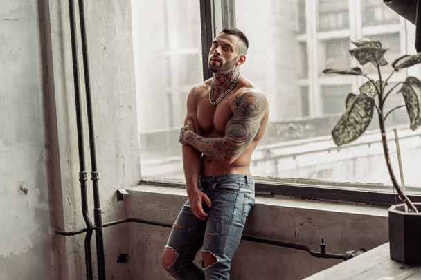 Fashion portrait of Sexy naked male model with tattoo and a black beard standing in hot pose on near the window. Loft room interior with grey concrete wall. Professional Studio image - Zdjęcie, obraz