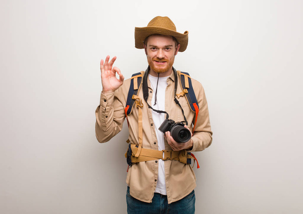 Young redhead explorer man cheerful and confident doing ok gesture. Holding a camera. - Photo, Image