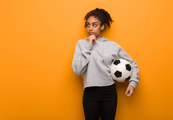 Молодая фитнес черная женщина сомневается и смущается. Холдинг футбольный мяч
. - Фото, изображение