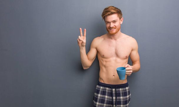 Young shirtless redhead man showing number two. He is holding a coffee mug. - Foto, afbeelding