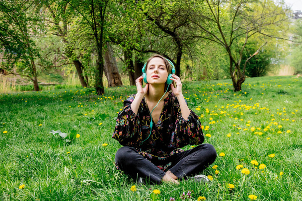 公園で緑のタンポポの牧草地にヘッドフォンを持つ若い女性.  - 写真・画像