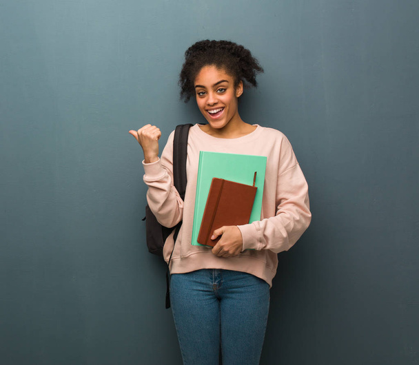 Nuori opiskelija musta nainen hymyilee ja osoittaa sivuun. Hänellä on kirjoja.
. - Valokuva, kuva