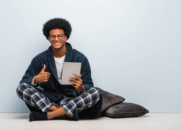 Giovane uomo nero seduto sulla sua casa e tenendo il tablet sorridente e alzando il pollice verso l'alto
 - Foto, immagini