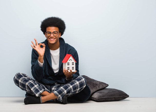 Joven hombre negro sosteniendo una casa modelo sentado en el suelo alegre y seguro haciendo buen gesto
 - Foto, imagen