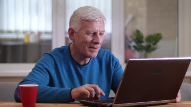 Closeup shoot of aged caucasian man having a video call on the laptop smiling happily indoors in a cozy apartment - Кадры, видео