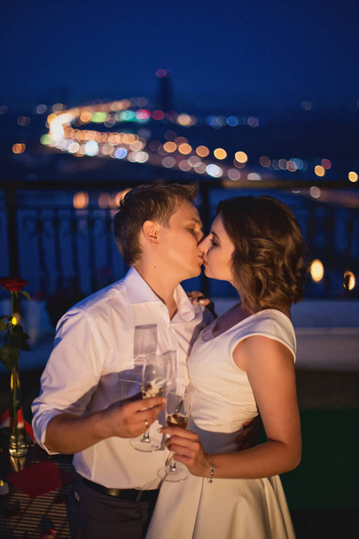 young couple of lovers celebrating love date and standing with wine glasses on bridge on night city view background - Foto, immagini