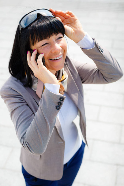 Smile Brunette business woman in street speaks by cell mobile phone and put hand in pocket with sunglasses on head, Positive human emotion, facial expression, spring mood - Photo, Image