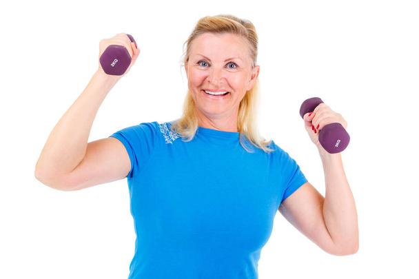 Pensioner woman sport. Old senior woman is engaged in inside sports. Woman looking at camera, holding violet dumbbells and cheerfully smiling while doing sport exercises. isolated on white background - Photo, Image