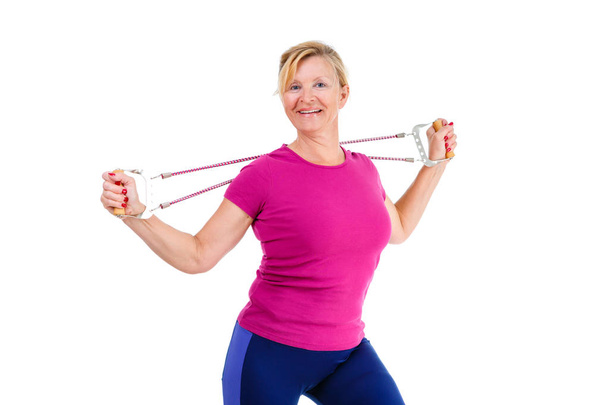 Mujer rubia mayor feliz y sonriente vestida de color camisa Marsala haciendo ejercicios de fitness con expansor una banda de resistencia, aislada sobre fondo blanco, emociones humanas positivas
 - Foto, Imagen