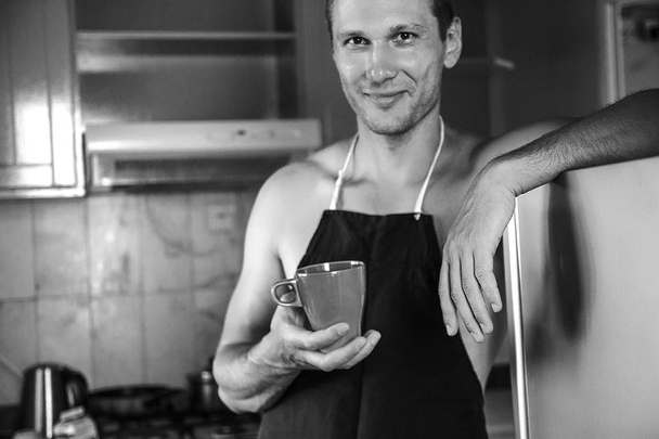 Улыбающийся мужчина в фартуке с чашкой кофе на кухне
. - Фото, изображение