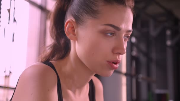 Closeup shoot of young fit athlete female sitting in the gym and looking forward breathing heavily with motivated facial expression indoors - Imágenes, Vídeo
