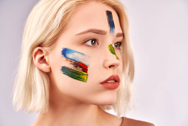 Face art concept. Pleasant woman with beautiful art makeup, has short blonde hair, poses against white background. Close up shot of beautiful female, looks thoughtfully at place of your advertising - Foto, afbeelding