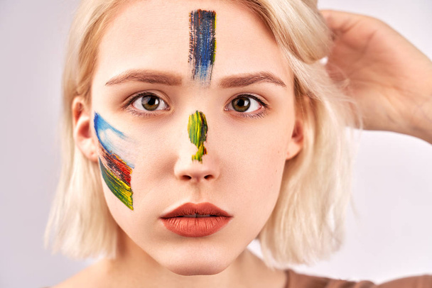 People, beauty and creative. Pleasant looking woman with beautiful art makeup on face, has short blonde hair, poses against white studio background. Lovely female has paint strokes on cheeks - Φωτογραφία, εικόνα