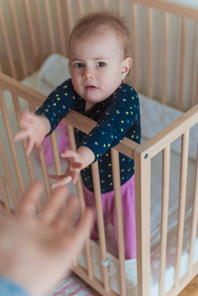 Yeni yürümeye başlayan bir kız bebek yatağında dururken ellerini annesine uzatır. Çocuk uyanıyor. Toddler kabuslar sorunu. Ayrılık korkusu. - Fotoğraf, Görsel