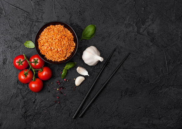 schwarze Tellerschüssel mit Reis mit Tomate und Basilikum und Knoblauch und Essstäbchen auf schwarzem Stein Hintergrund. Ansicht von oben. - Foto, Bild