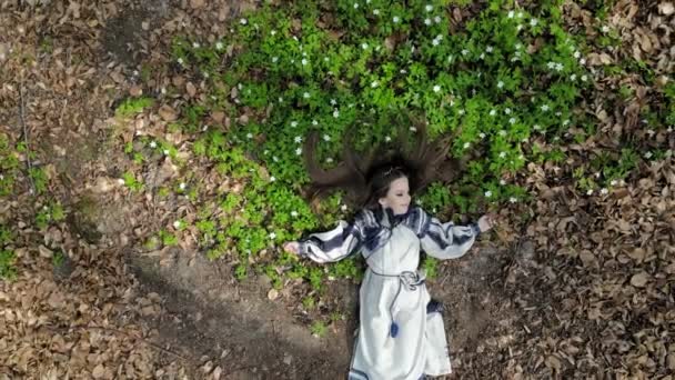Vista aérea no topo. Uma menina está deitada na grama com flores brancas com os olhos fechados. A câmera se afasta lentamente e se aproxima. Tiro de um drone
 - Filmagem, Vídeo