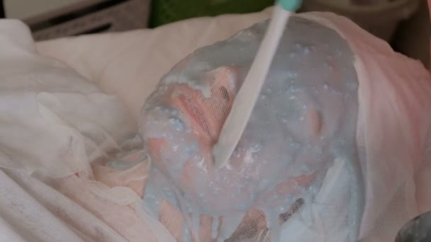 Het meisje met gaas op het gezicht ligt op de procedure in de schoonheidssalon. De cosmetoloog past een verjongende blauwe masker toe. Close-up. - Video