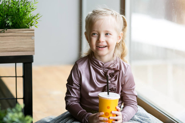 A small child drinks juice from a paper cup with a straw sitting by the window. Little girl with a paper cup of juice. - Photo, Image