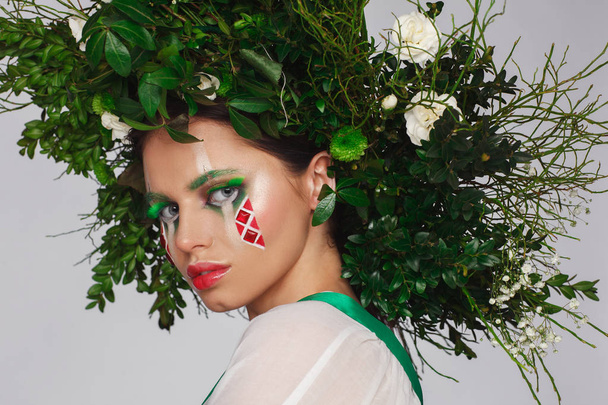 Portrait of a beautiful woman in a white dress with a green ribbon and with branches on her head. Creative makeup - Foto, imagen