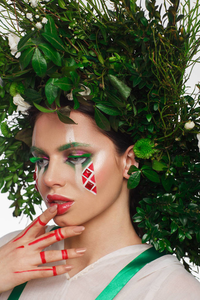 Retrato de una hermosa mujer en un vestido blanco con una cinta verde y con ramas en la cabeza. Maquillaje creativo
 - Foto, imagen