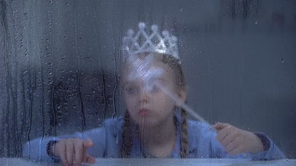 Menina perturbada na coroa com pau mágico sentado sozinho atrás da janela chuvosa
 - Filmagem, Vídeo