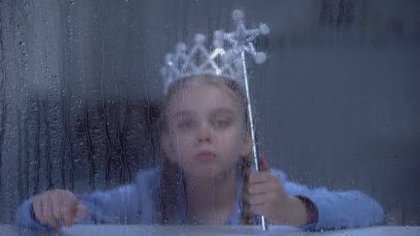 Μοναχική μικρή πριγκίπισσα με στέμμα με μαγικό ραβδί κάθεται πίσω από βροχερό παράθυρο - Πλάνα, βίντεο