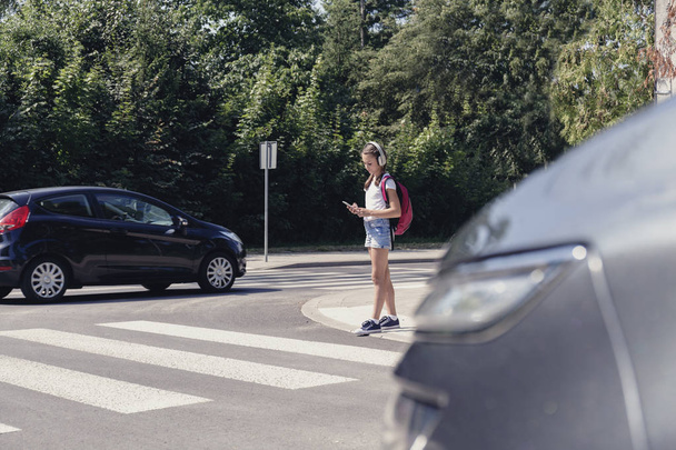 Schoolgirl with headphones and mobile phone on pedestrian crossing in front of a car - Foto, Bild