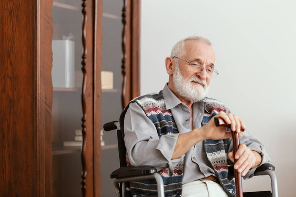 Elderly man sitting on a wheelchair and supporting himself with cane - Foto, Imagem