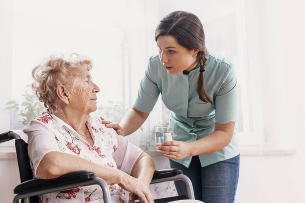 Senior grandmother on wheelchair, supporting nurse giving her glass of water - Foto, imagen