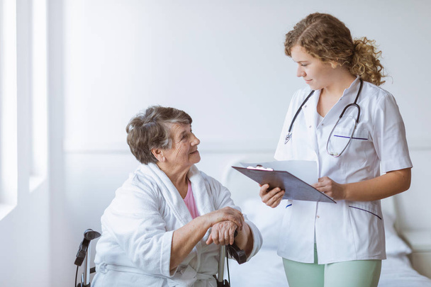 Young intern doctor with pad and stethoscope and elderly grandmother with cane - Photo, image