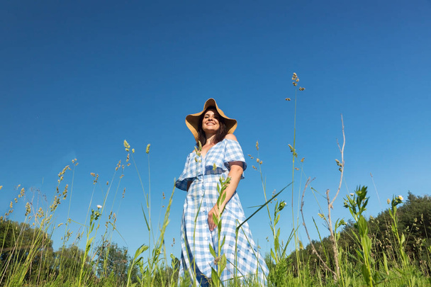 A woman in a blue dress and a straw hat stands in the field and  - Foto, immagini