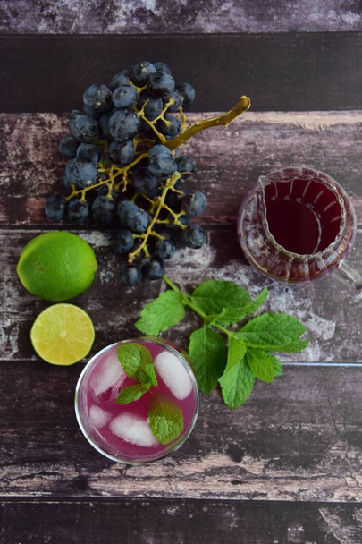 Refrescante bebida de uva roja en vaso con cubitos de hielo decorado con hojas de menta. Fruta de uva en el fondo - Foto, imagen