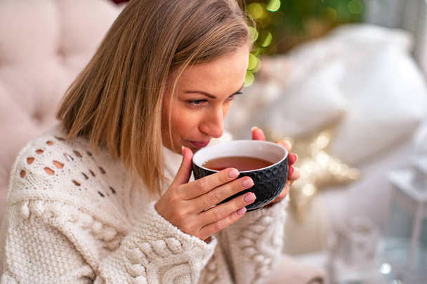 blonde fille dans un pull avec une tasse de thé assis sur le lit
 - Photo, image