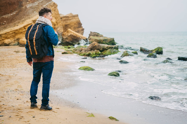 Traveler with a backpack standing near a rock against a beautiful sea with waves, a stylish hipster boy posing near a calm ocean during a wonderful journey around the world. Shoot from the back - Foto, Imagen