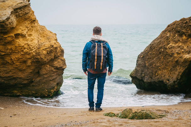 Traveler with a backpack standing near a rock against a beautiful sea with waves, a stylish hipster boy posing near a calm ocean during a wonderful journey around the world. Shoot from the back - Foto, immagini