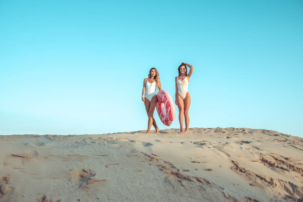 Beautiful girls white swimsuit bodysuit. Walk in sand. Sunbathe resting nature. Girlfriends sister. Concept getaway nature. Fashion lifestyle is young. They go swimming in hands circle swimming, toy. - Photo, Image