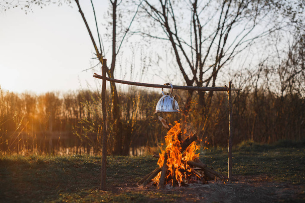 Teapot with tea hangs over the fire on the riverbank sunset - Fotoğraf, Görsel