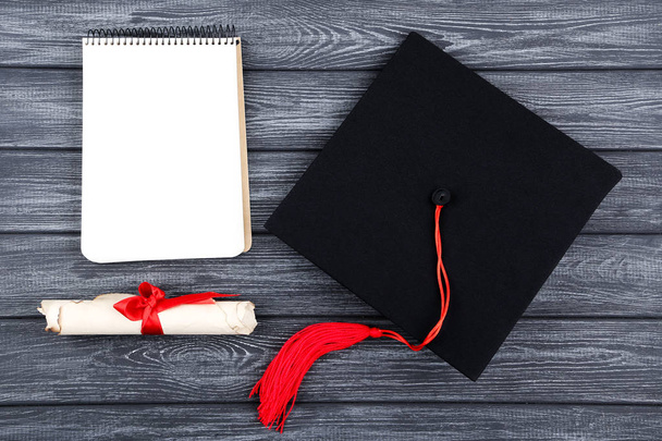 Graduation cap with diploma and notepad on black wooden table - Φωτογραφία, εικόνα
