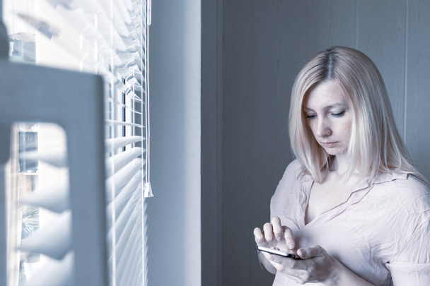 Sad apathetic lonely woman standing near the window and texting messages in smartphone at home or hotel, divorce, depression and apathy concept - Foto, afbeelding